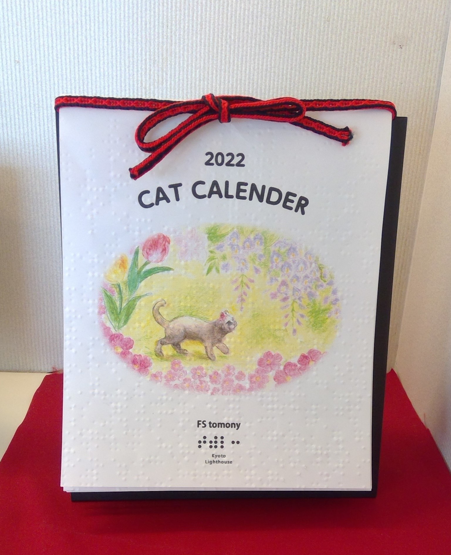 点字用紙に印刷された「2022トモニーカレンダー」の表紙には桜の下を歩く猫の絵。赤い紐で結んであります。